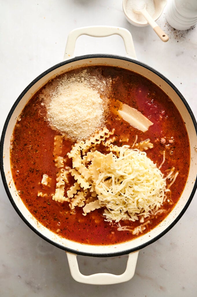 Tomates triturados, fideos de lasaña rotos, mozzarella rallada y parmesano añadidos a la olla.