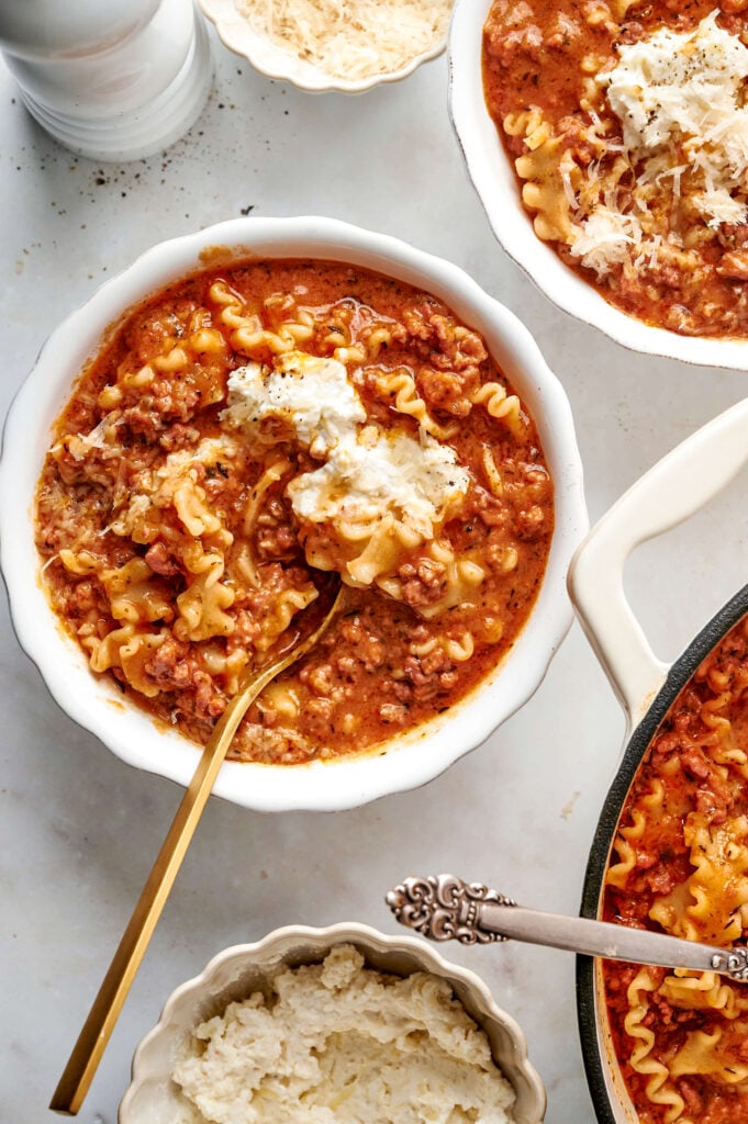 Un tazón de sopa de lasaña cremosa con fideos ondulados, rico caldo de tomate, carne molida y una cucharada de queso ricotta, servido con una cuchara dorada.
