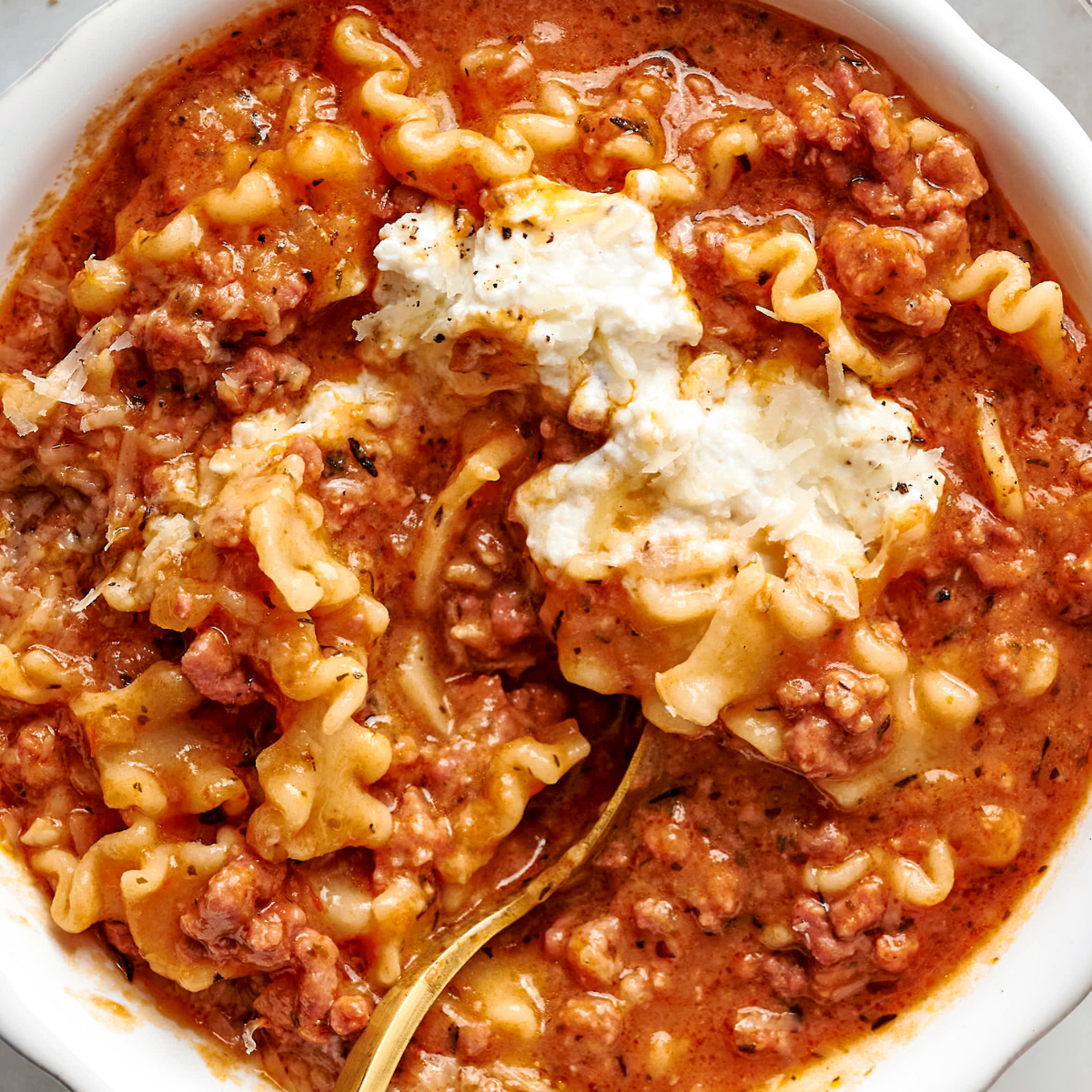 Acercamiento de sopa cremosa de lasaña en un tazón, coronada con una cucharada de queso ricotta y servida con una cuchara dorada.