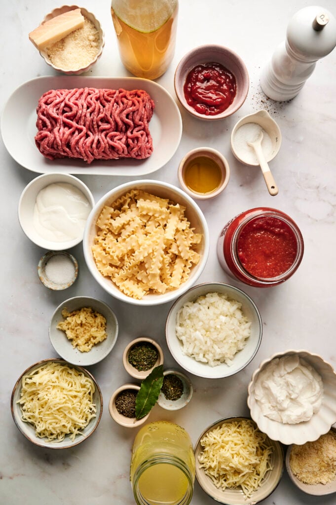 Ingredientes para la sopa de lasaña dispuestos sobre una superficie blanca, incluyendo pasta, carne molida, pasta de tomate, caldo y queso.