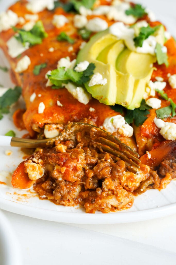 Acercamiento de una enchilada de carne con queso cubierta con rodajas de aguacate, cilantro y queso desmenuzado, con un bocado sacado para mostrar el relleno con salsa