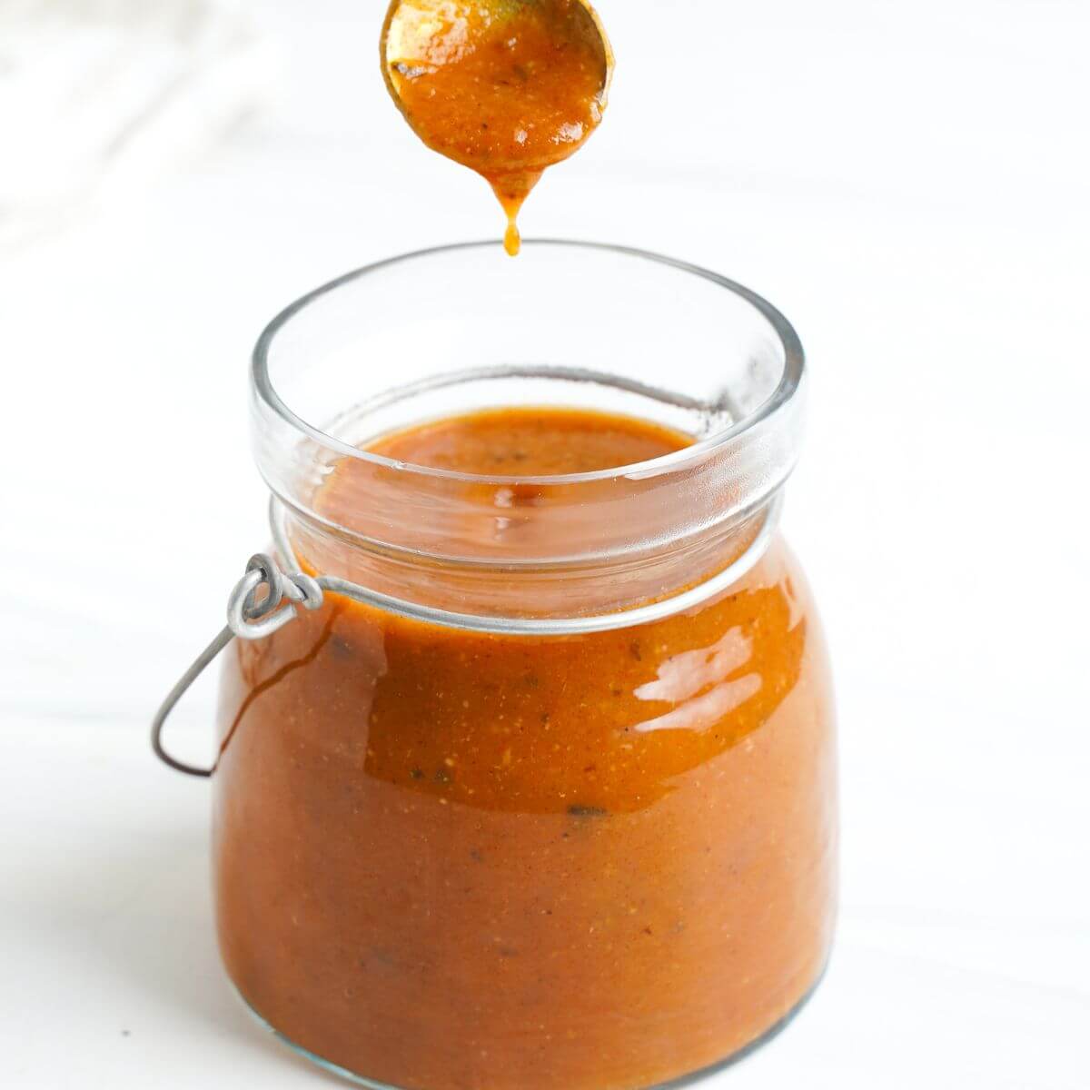 Un frasco de vidrio lleno de salsa para enchiladas con una cuchara goteando la salsa, hecha de tomates asados a la leña y especias, contra un fondo blanco limpio.