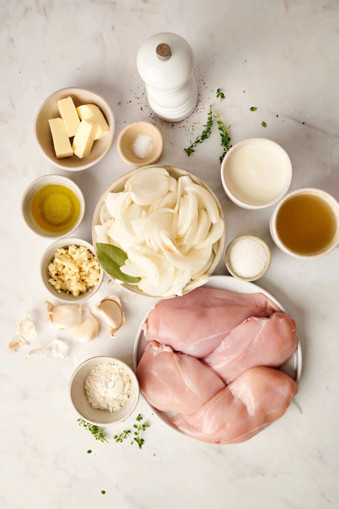 Vista aérea de los ingredientes para Pollo Cremoso con Cebolla Francesa, incluyendo pechugas de pollo crudas, cebollas dulces en rodajas, ajo, tomillo fresco, mantequilla, aceite de oliva, crema espesa, caldo de pollo, harina, hoja de laurel, sal y pimienta sobre una superficie blanca.