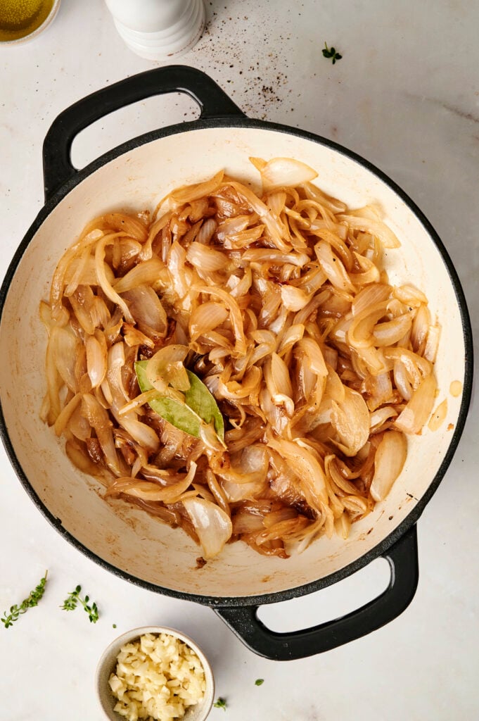 Cebollas profundamente caramelizadas en una sartén con hoja de laurel, creando una base rica para el pollo cremoso con cebolla francesa.