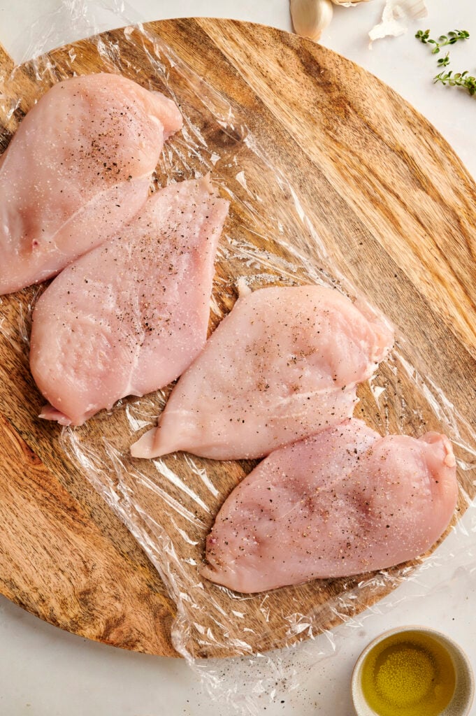 Pechugas de pollo crudas colocadas sobre una tabla de cortar de madera, sazonadas con sal y pimienta, sobre envoltura de plástico.