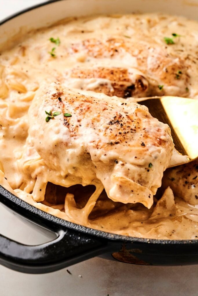 Primer plano de una pechuga de pollo dorada siendo levantada de una sartén llena de salsa cremosa de cebolla francesa y cebollas caramelizadas.