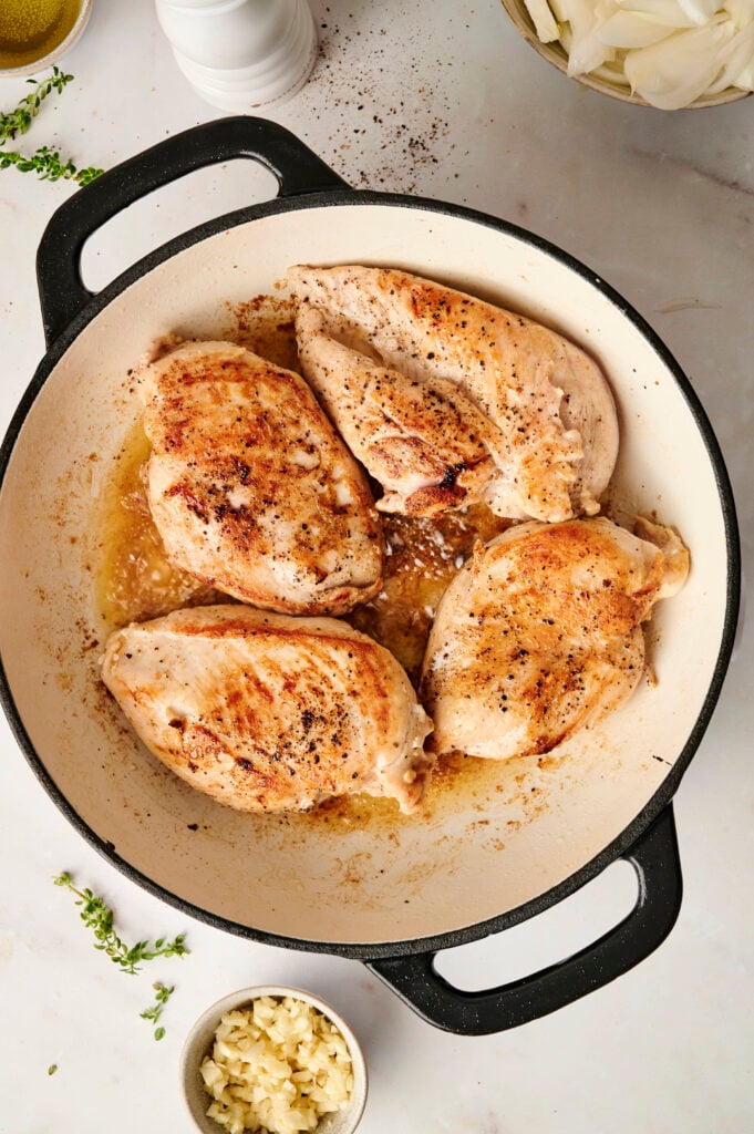 Pechugas de pollo doradas en una sartén blanca, listas para ser cubiertas con cebollas caramelizadas y salsa de crema.