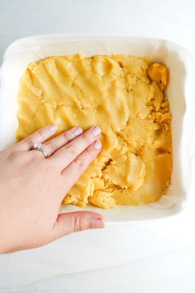 Mano presionando la masa de galleta de mantequilla uniformemente en un molde forrado con papel pergamino para las barras de limón.