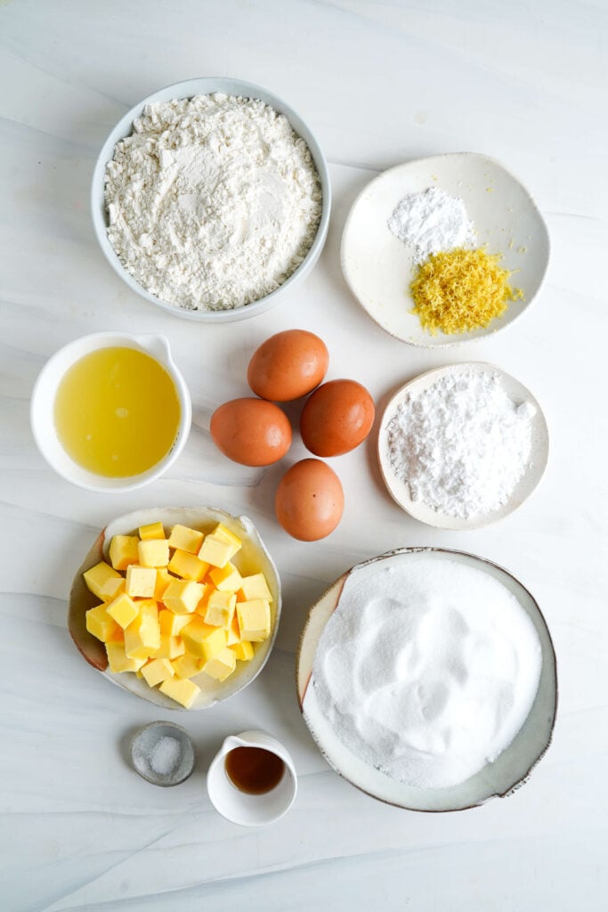 Ingredientes para la receta de barras de limón exhibidos sobre una superficie blanca, incluyendo harina, azúcar granulada, mantequilla, huevos, jugo de limón, ralladura de limón, azúcar en polvo, sal, extracto de vainilla, y polvo de hornear.