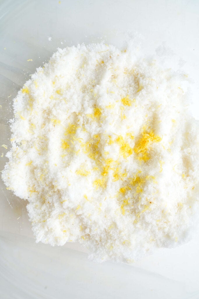 Azúcar granulada mezclada con ralladura de limón fresca en un tazón, mostrando el paso donde se liberan los aceites cítricos para realzar el sabor de la barra de limón.