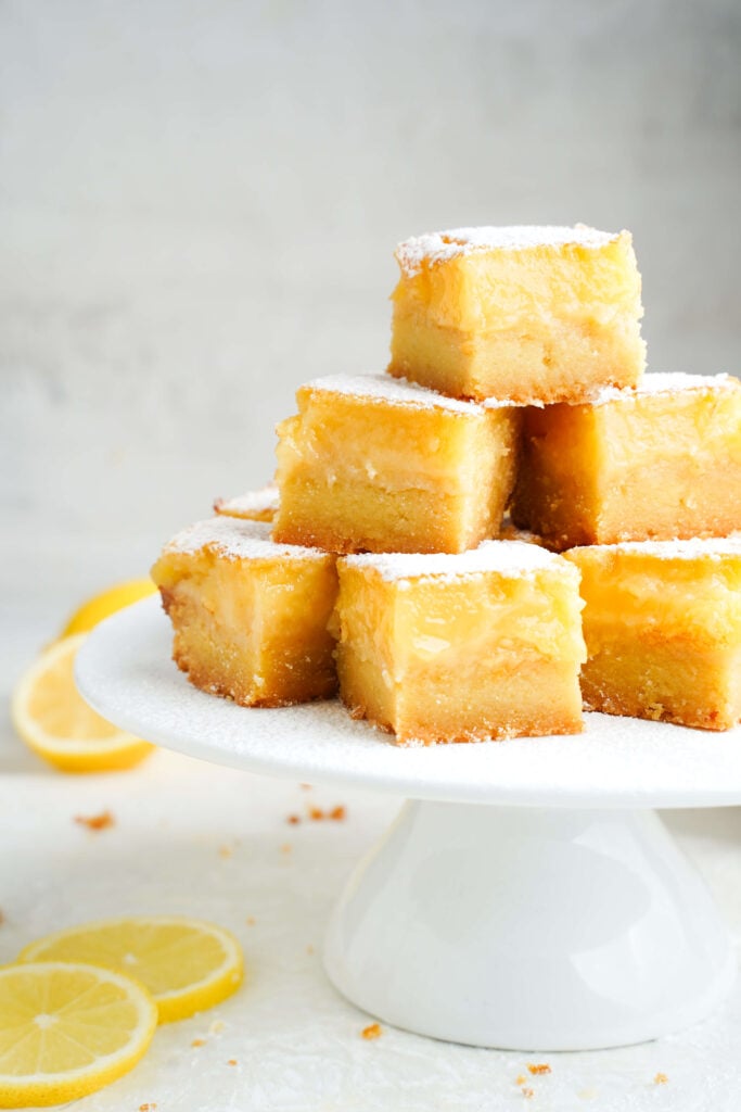 Barras de limón apiladas en un soporte para pasteles blanco, espolvoreadas con azúcar en polvo y rodeadas de frescas rodajas de limón.