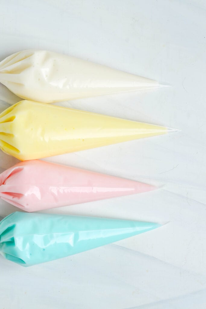 Cuatro mangas pasteleras llenas de glaseado de Pascua en colores pastel en blanco, amarillo, rosa y azul, listas para decorar galletas.