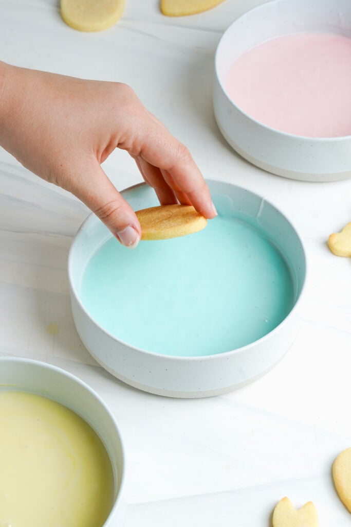 Mano sumergiendo una galleta de azúcar de Pascua en un tazón de glaseado azul pastel para decorar.