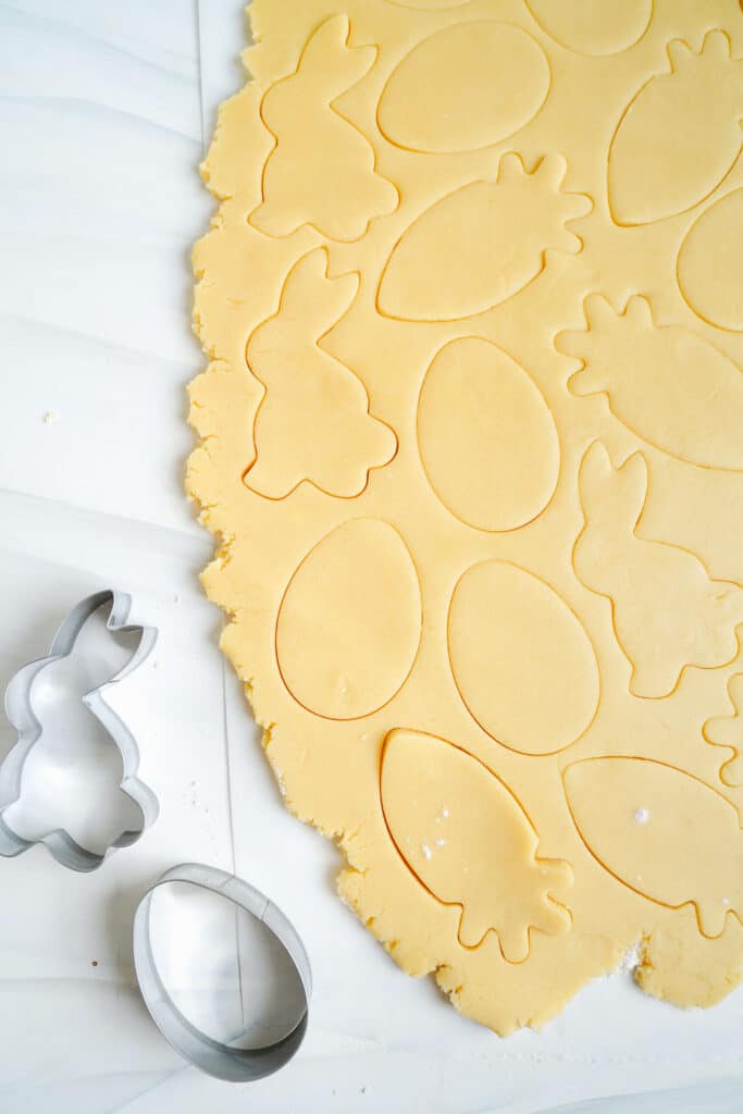 Masa de galleta de azúcar estirada y cortada en formas de conejos, huevos y zanahorias con cortadores de galletas de metal al lado.