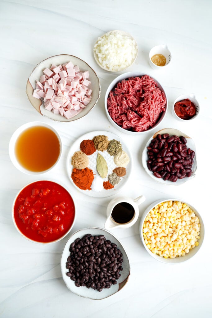 Una variedad de ingredientes para chili con carne, que incluyen carne molida, bacon, frijoles negros, frijoles rojos, tomates asados al fuego, maíz y una mezcla de especias.