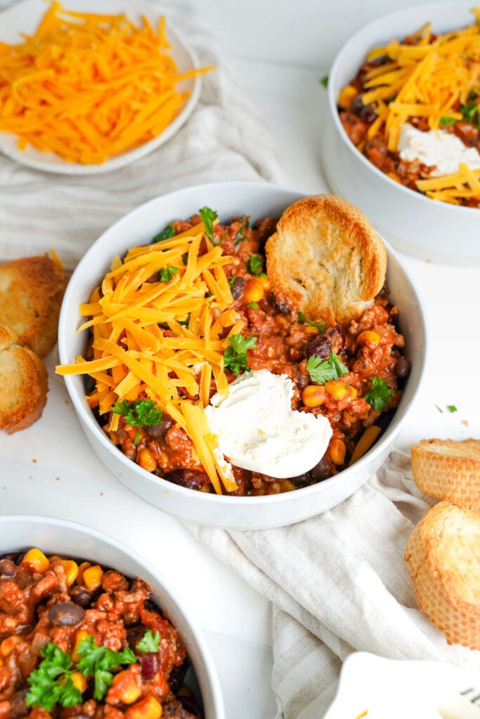 Chili con carne en un tazón, coronado con queso cheddar rallado, crema agria y perejil fresco, con rodajas de baguette tostadas al lado.