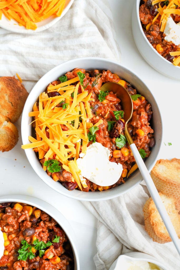 Mejor chili con carne servido en tazones con queso cheddar rallado, crema agria y rodajas de baguette tostadas para mojar. Una cuchara está colocada en uno de los tazones, lista para disfrutar.