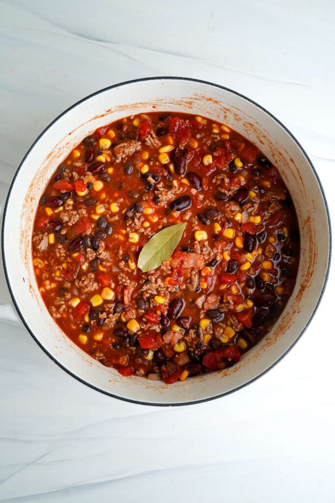 Chili con carne cociendo a fuego lento en una olla de hierro fundido con frijoles, maíz, tomates asados al fuego y una hoja de laurel.
