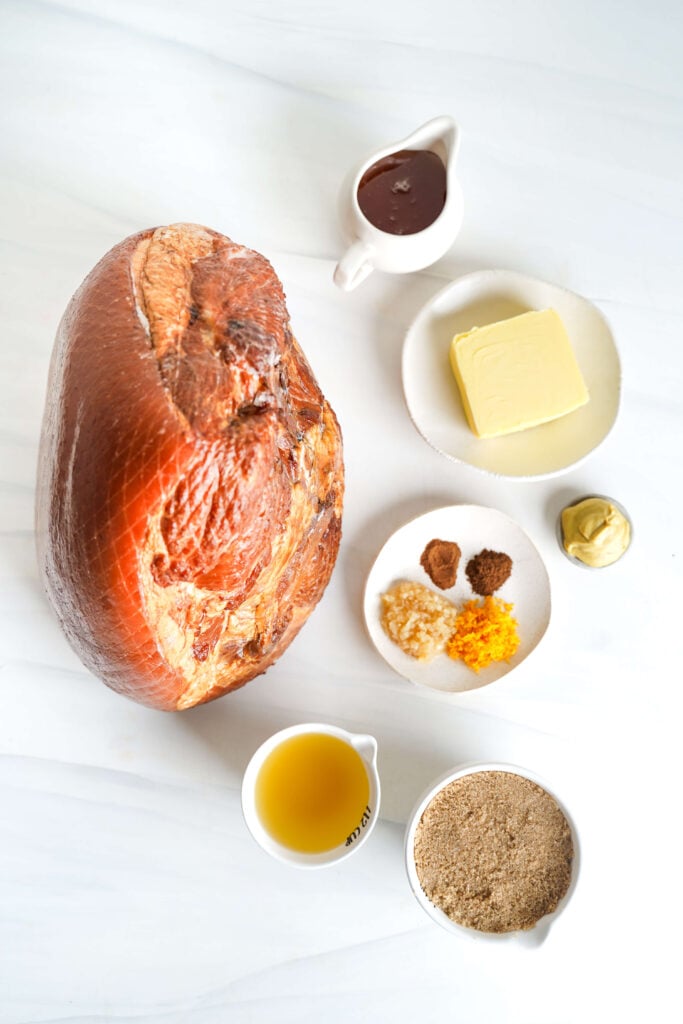 Vista desde arriba de ingredientes para una receta de jamón glaseado con azúcar moreno, incluyendo jamón con hueso, mantequilla, azúcar moreno, miel, mostaza dijon, ajo, especias, ralladura de naranja y caldo de pollo