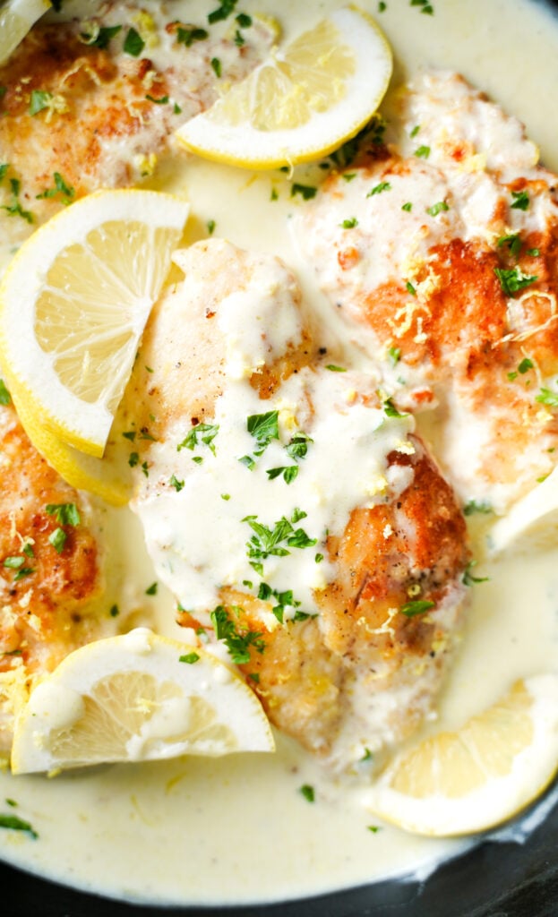 Primer plano de pechugas de pollo cremoso al limón en una rica y mantequilla salsa, adornadas con perejil fresco y rodajas de limón en una sartén negra.