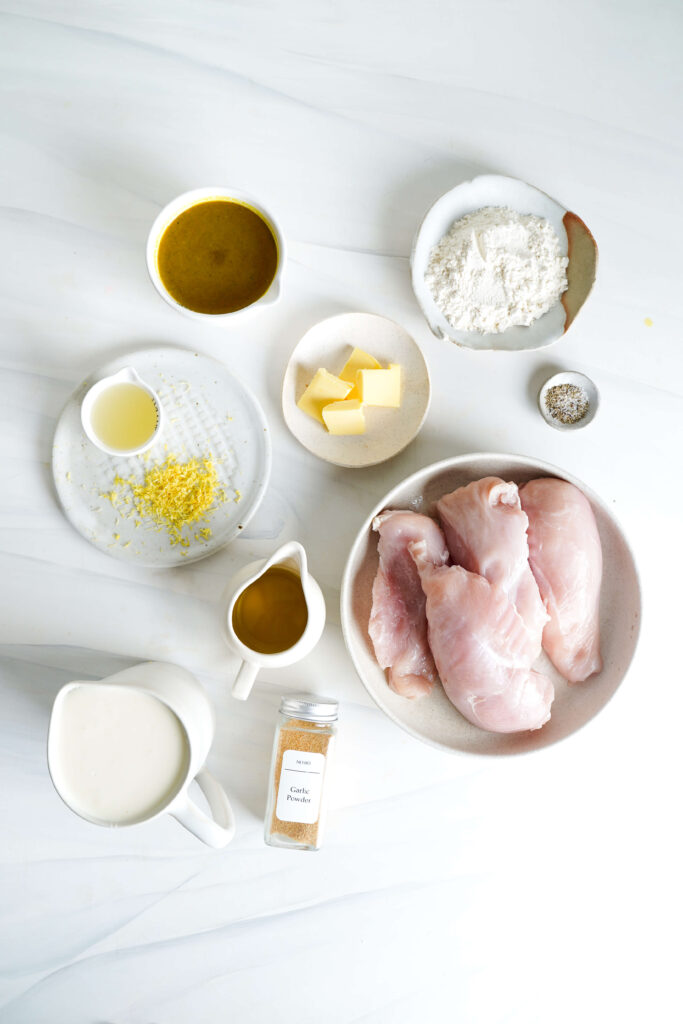 Presentación plana de ingredientes para pasta de pollo cremoso al limón, incluyendo pechugas de pollo crudas, crema espesa, mantequilla, harina, caldo de pollo, polvo de ajo, aceite de oliva, jugo de limón y ralladura, sal y pimienta sobre una superficie blanca.