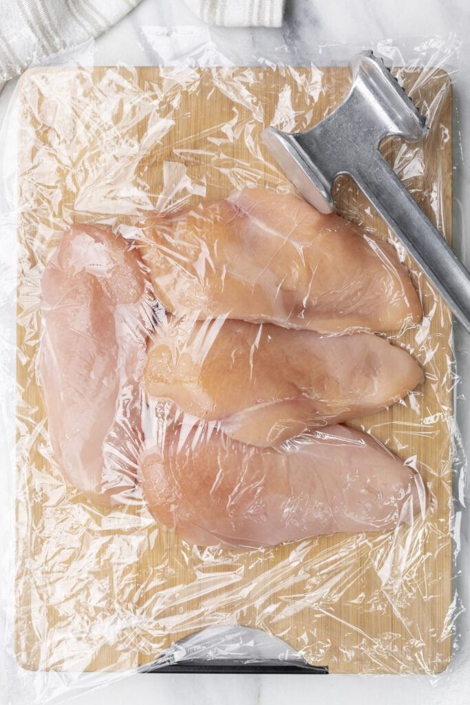 Pechugas de pollo aplanadas sobre una tabla de cortar de madera con el mazo de carne a un lado.