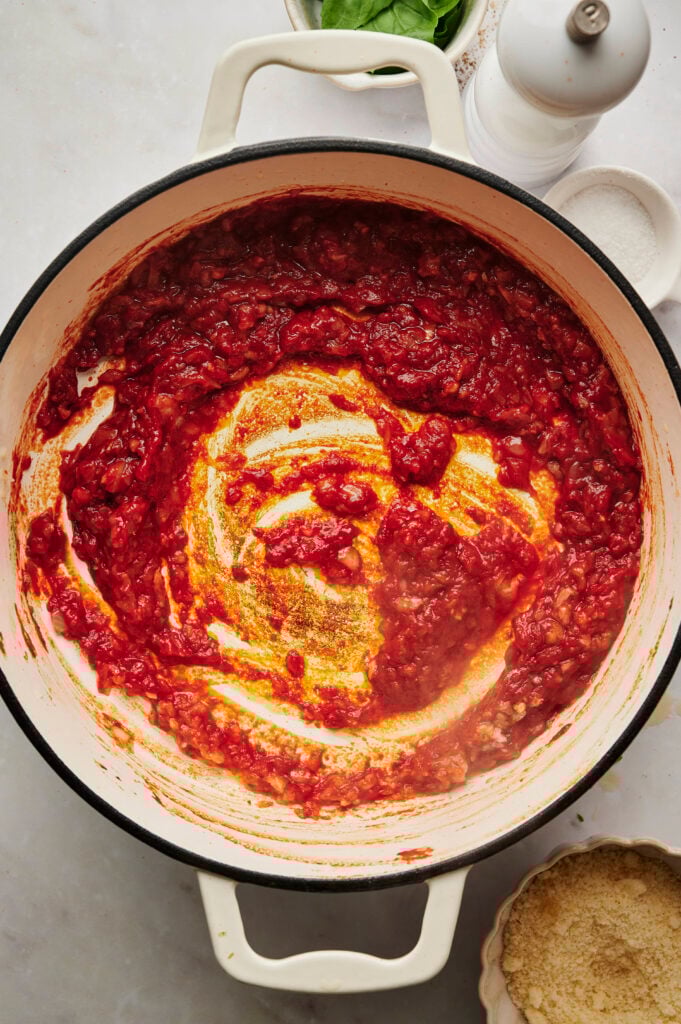 Pasta de tomate roja rica cocinada con aromáticos hasta oscurecer, formando la base de sabor con trozos marrones visibles en el fondo de la sartén blanca