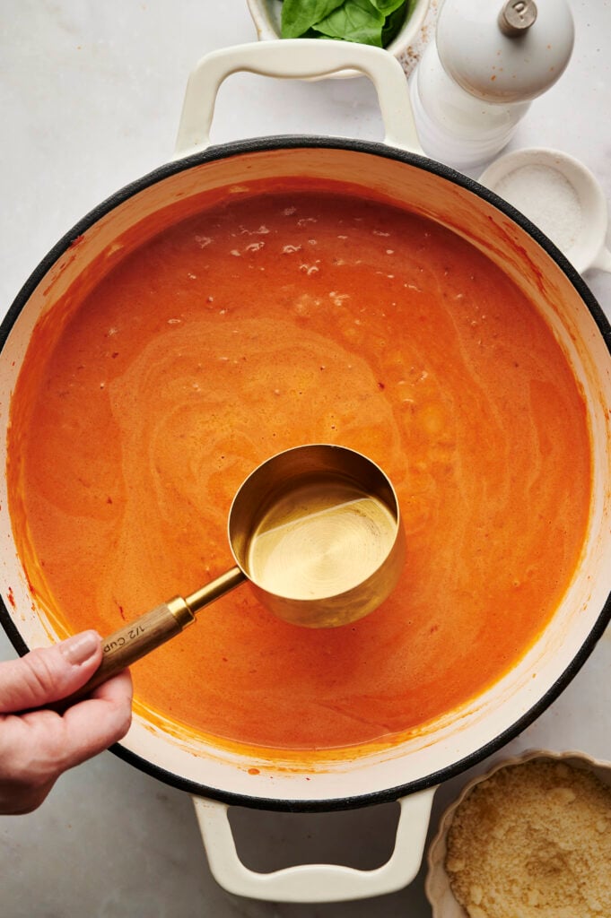 Mano sosteniendo una taza medidora de agua de pasta reservada sobre una sartén de salsa de vodka cremosa, lista para mezclar