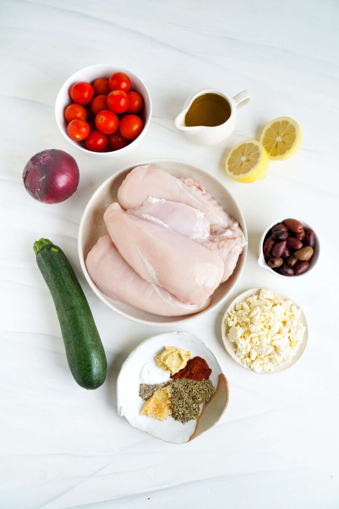Vista aérea de los ingredientes para pollo griego en bandeja, incluyendo pechugas de pollo crudas, tomates cherry, calabacín, cebolla roja, aceitunas Kalamata, feta desmenuzado, mitades de limón, aceite de oliva y un tazón de especias y mostaza de Dijon