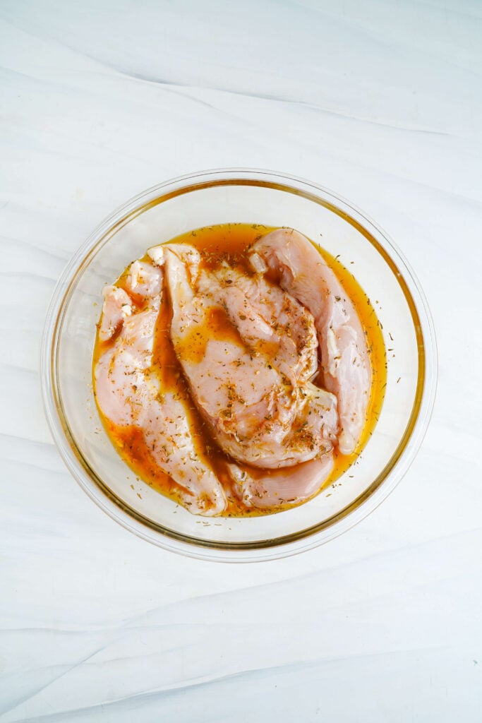 Pechugas de pollo crudas en un tazón de vidrio marinando en una mezcla de limón y hierbas para pollo griego en bandeja