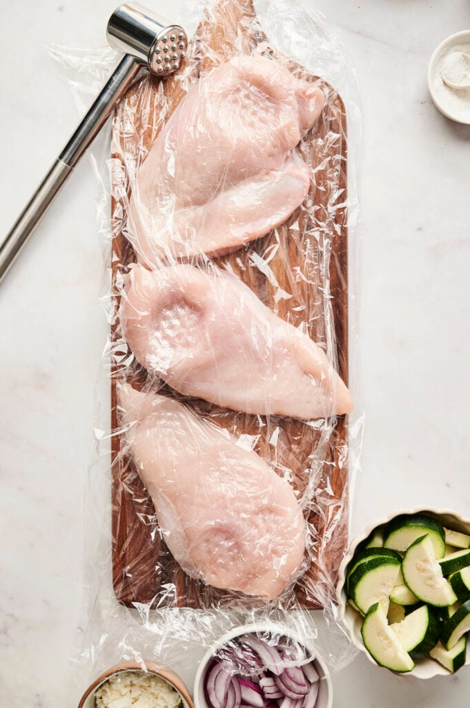 Pechugas de pollo crudas entre papel film en una tabla de cortar con un mazo de carne cerca