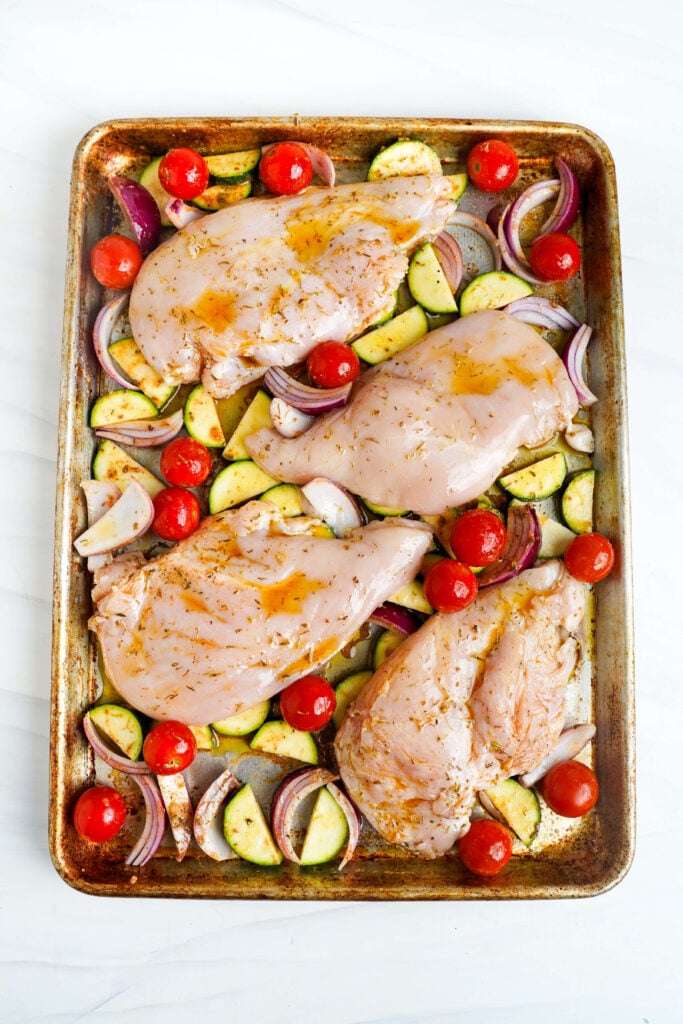 Pechugas de pollo crudas marinadas dispuestas en una bandeja con calabacín, tomates cherry y cebolla roja