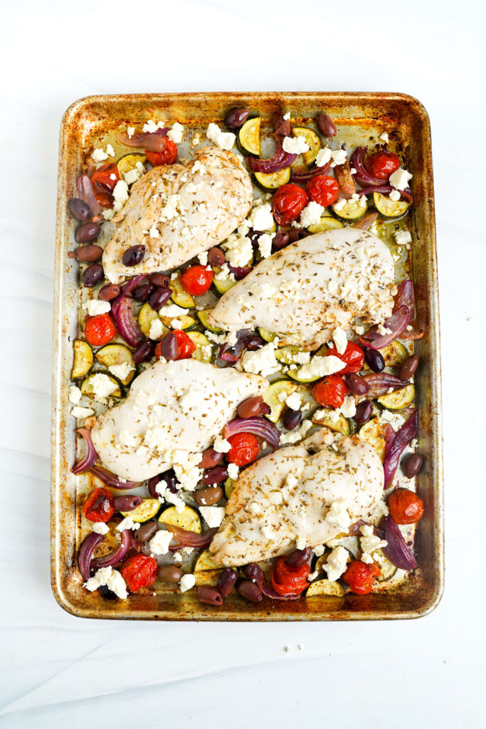 Pechugas de pollo en una bandeja con verduras, aceitunas y feta desmenuzado antes de regresar al horno