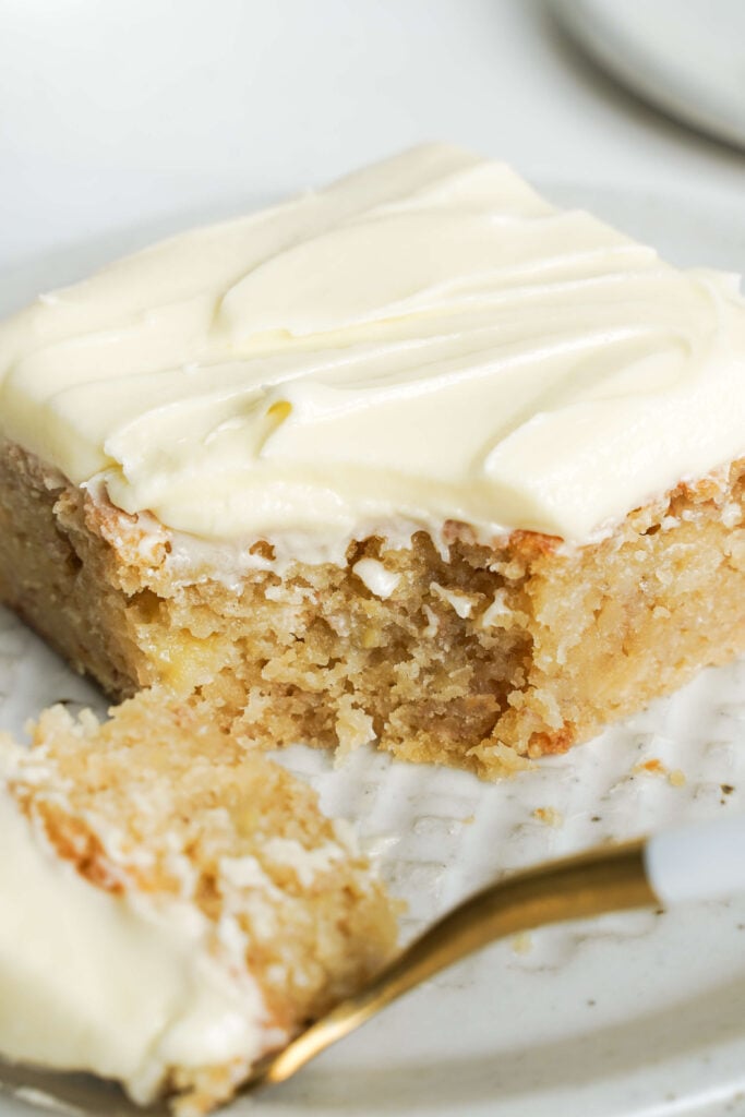 Primer plano de una rebanada de pastel de plátano húmedo con glaseado de queso crema, mostrando un bocado con un tenedor dorado y blanco