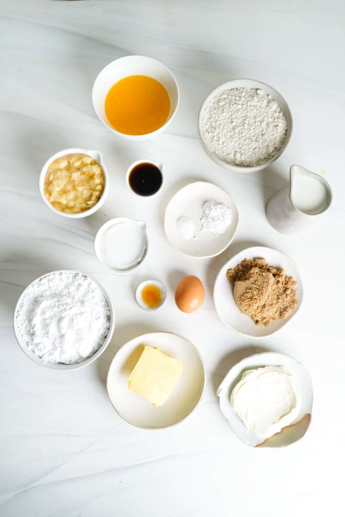 Vista desde arriba de los ingredientes para pastel de plátano con glaseado de queso crema, incluyendo plátanos machacados, harina, mantequilla derretida, leche, azúcar moreno, azúcar granulada, huevo, vainilla, polvo de hornear, sal, azúcar glas, queso crema y mantequilla organizados en una encimera blanca.