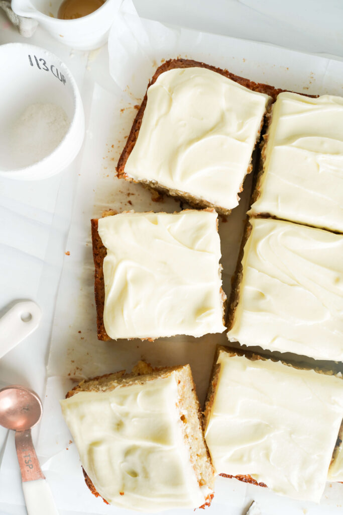 Vista desde arriba del pastel de plátano rebanado con glaseado de queso crema sobre papel pergamino, decorado con cucharas medidoras blancas y una taza de cerámica