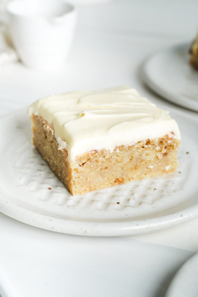 Porción de pastel de plátano con glaseado de queso crema en un plato blanco, mostrando su textura húmeda y su gruesa cobertura cremosa