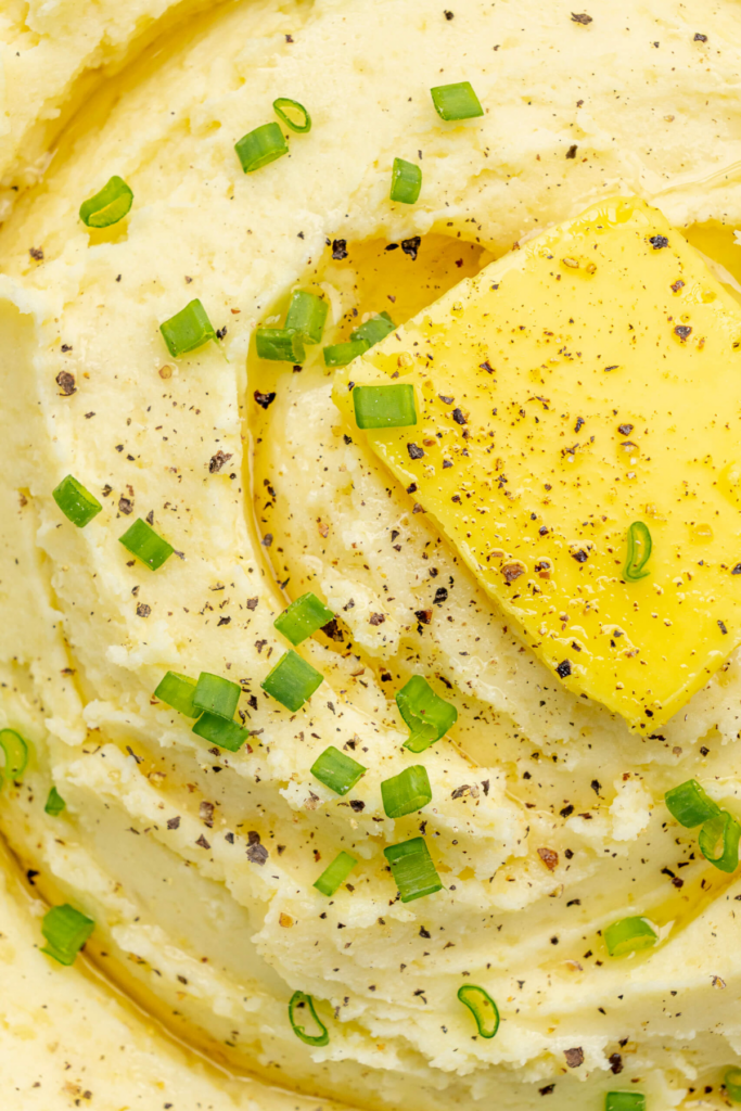 Primer plano del puré cremoso de patatas cubierto con un trozo de mantequilla, pimienta negra recién molida y cebollinos picados.