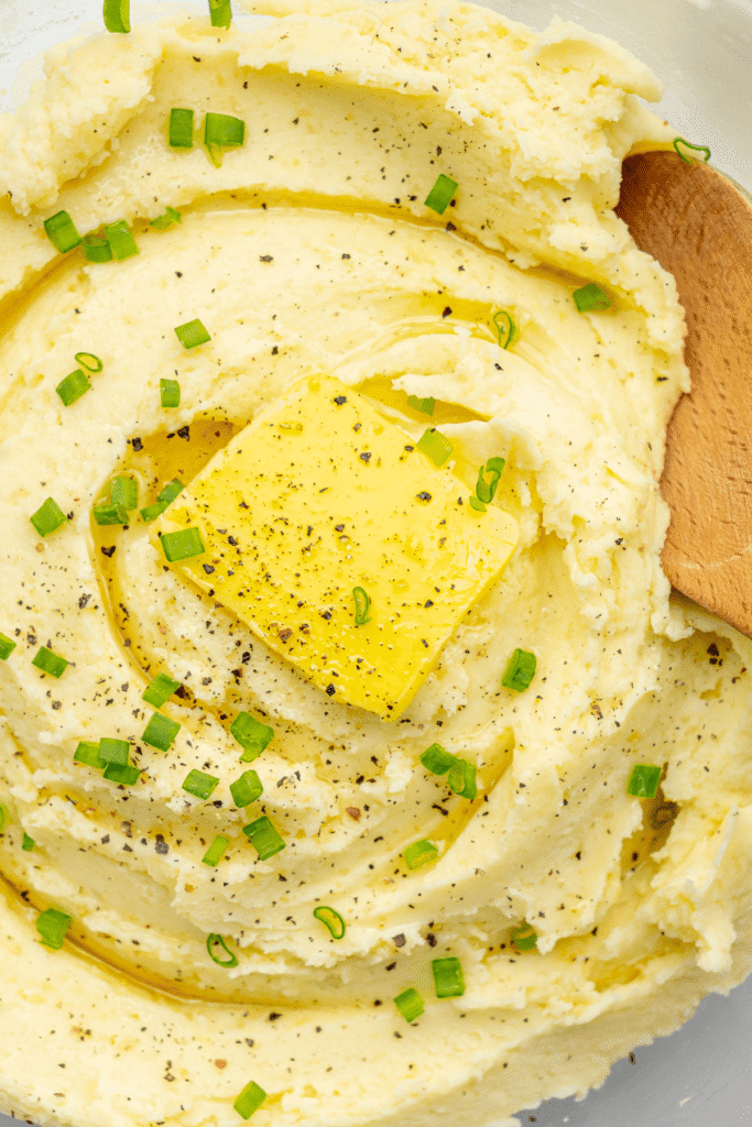 Primer plano del puré de patatas cubierto con un trozo de mantequilla, pimienta negra recién molida y cebollinos picados, listo para servir.