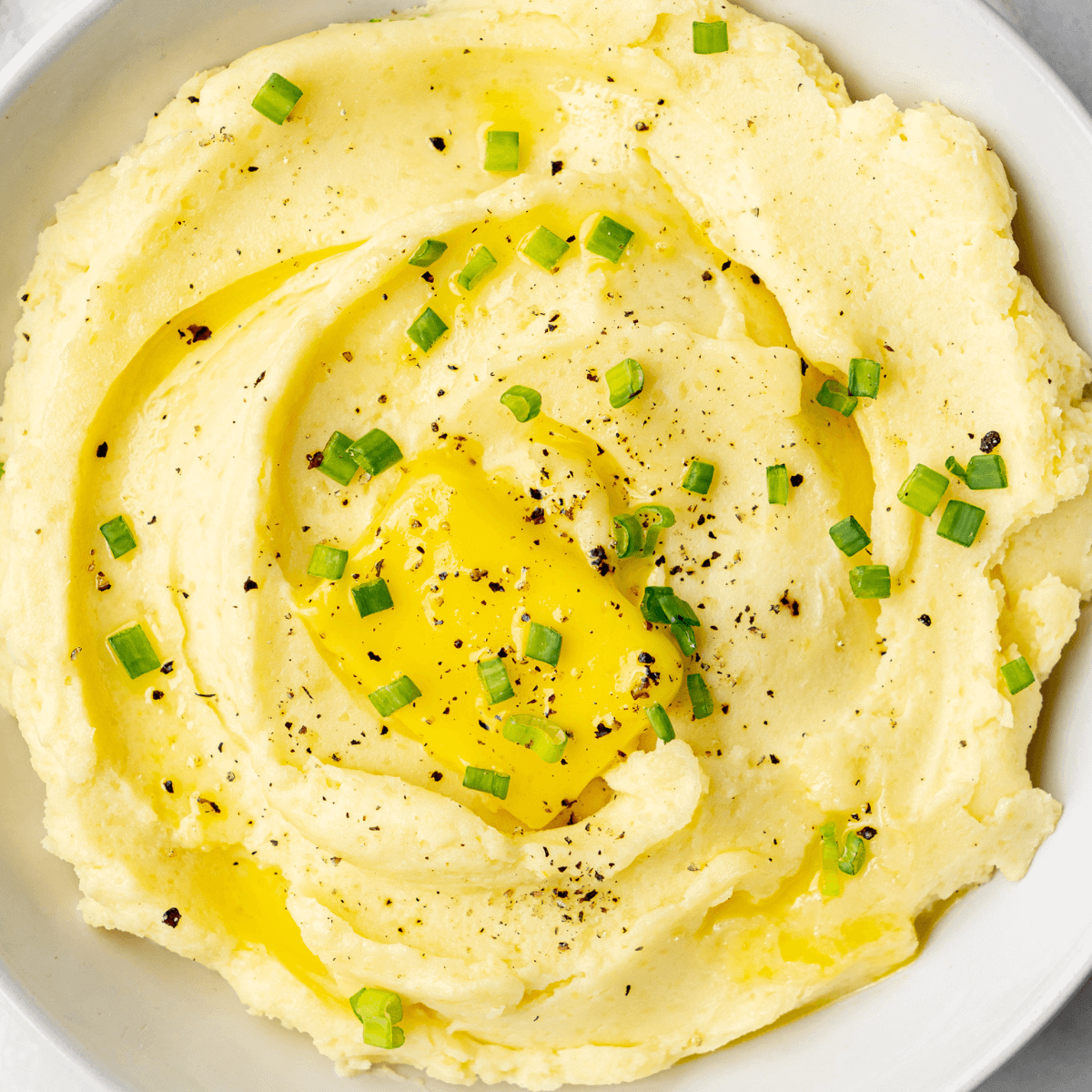 Primer plano del puré cremoso de patatas cubierto con un trozo de mantequilla, espolvoreado con pimienta negra recién molida y cebollinos picados.