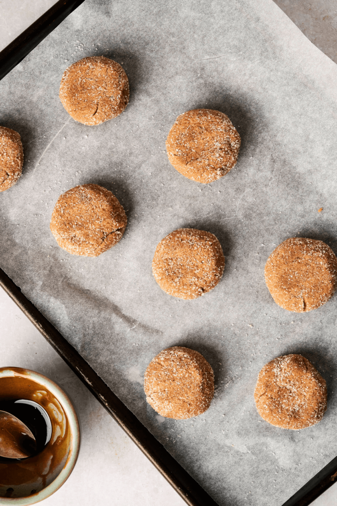 Bolas de masa de galletas de melaza sin hornear, cubiertas de azúcar y ligeramente aplanadas, organizadas en una bandeja para hornear forrada de papel pergamino. Un bol con melaza y una cuchara está cerca.