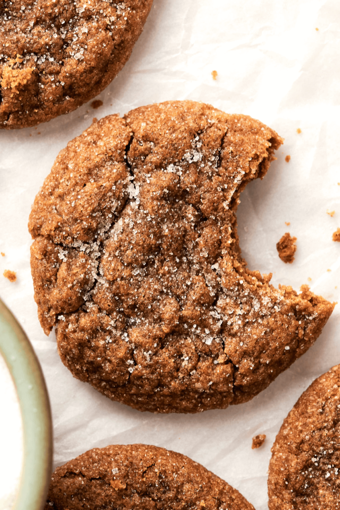 Primer plano de una galleta de jengibre y melaza con una parte superior agrietada cubierta de azúcar y un mordisco tomado, mostrando su interior suave. Otras galletas son visibles en papel pergamino en el fondo.