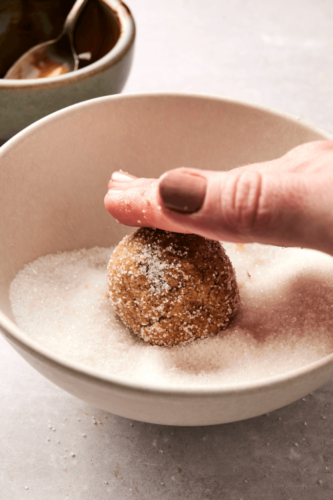 Una mano presionando una bola de masa de galleta de jengibre y melaza en un bol de azúcar granulada, preparándola para hornear. Un pequeño bol con una cuchara es visible en el fondo.