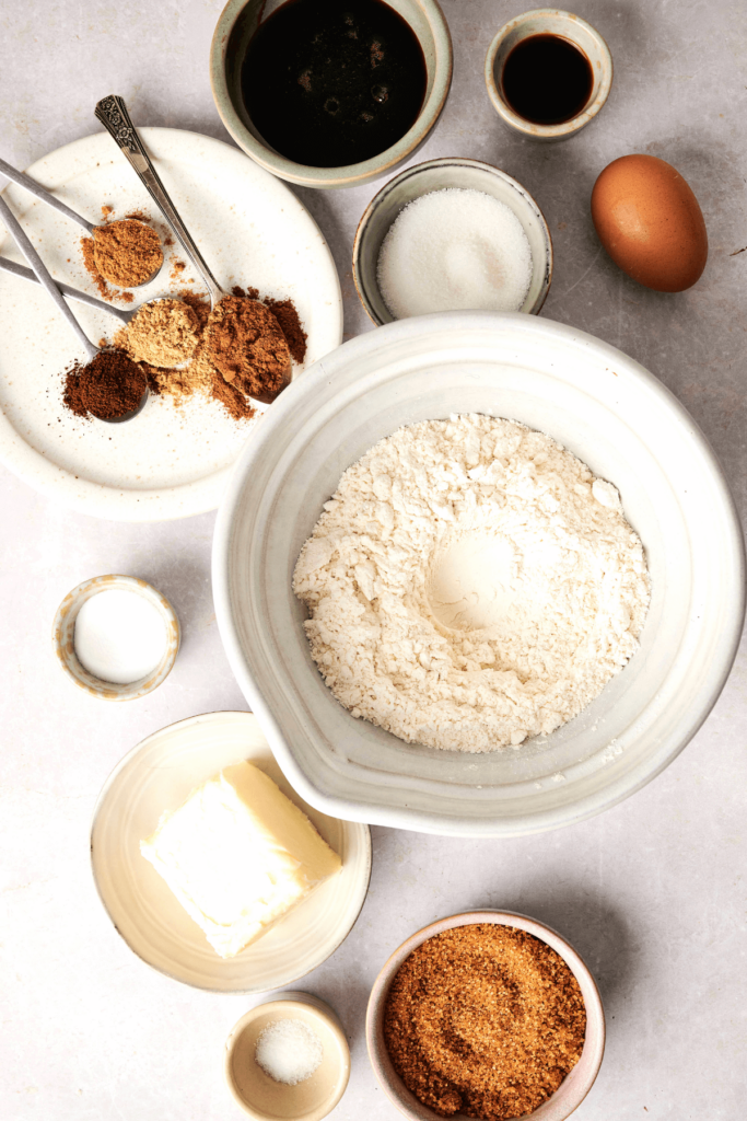 Distribución de ingredientes para galletas de jengibre y melaza, incluyendo un bol de harina, azúcar moreno, mantequilla sin sal, un huevo, melaza, extracto de vainilla, especias molidas (jengibre, canela, clavos, nuez moscada) y azúcar granulada.
