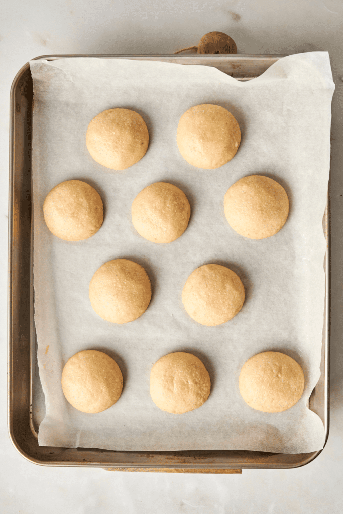 Galletas de Almendra en Forma de Nieve recién horneadas y doradas, enfriándose en una bandeja de hornear forrada de papel antes de ser cubiertas con azúcar en polvo.