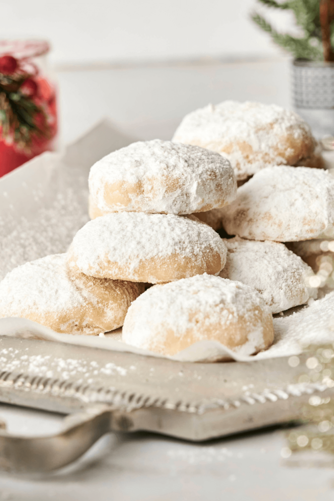 Un montón de Galletas de Almendra en Forma de Nieve cubiertas de azúcar en polvo en una bandeja para servir, rodeadas de decoraciones festivas, mostrando su suave y azucarada superficie.