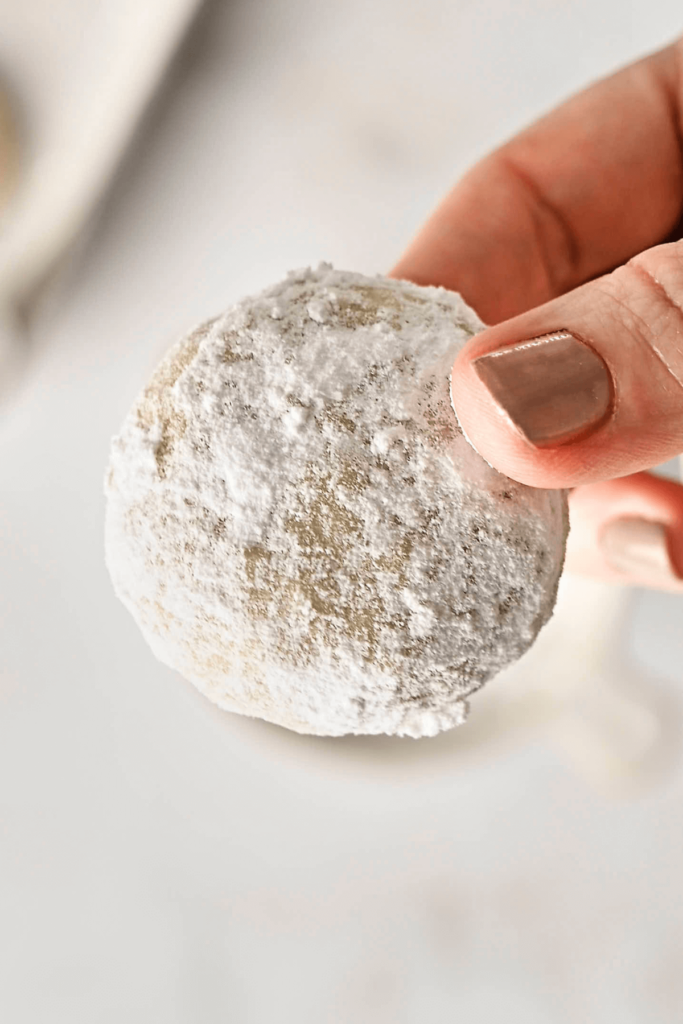 Una mano sosteniendo una Galleta de Almendra en Forma de Nieve recién cubierta de azúcar, mostrando su delicada y azucarada superficie.
