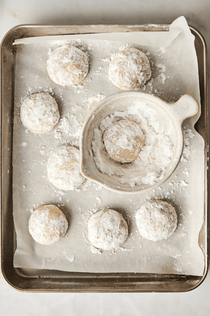 Galletas de Almendra en Forma de Nieve recién horneadas siendo cubiertas con azúcar en polvo, con una galleta descansando en un tazón de azúcar en polvo y otras en una bandeja de hornear forrada de papel.