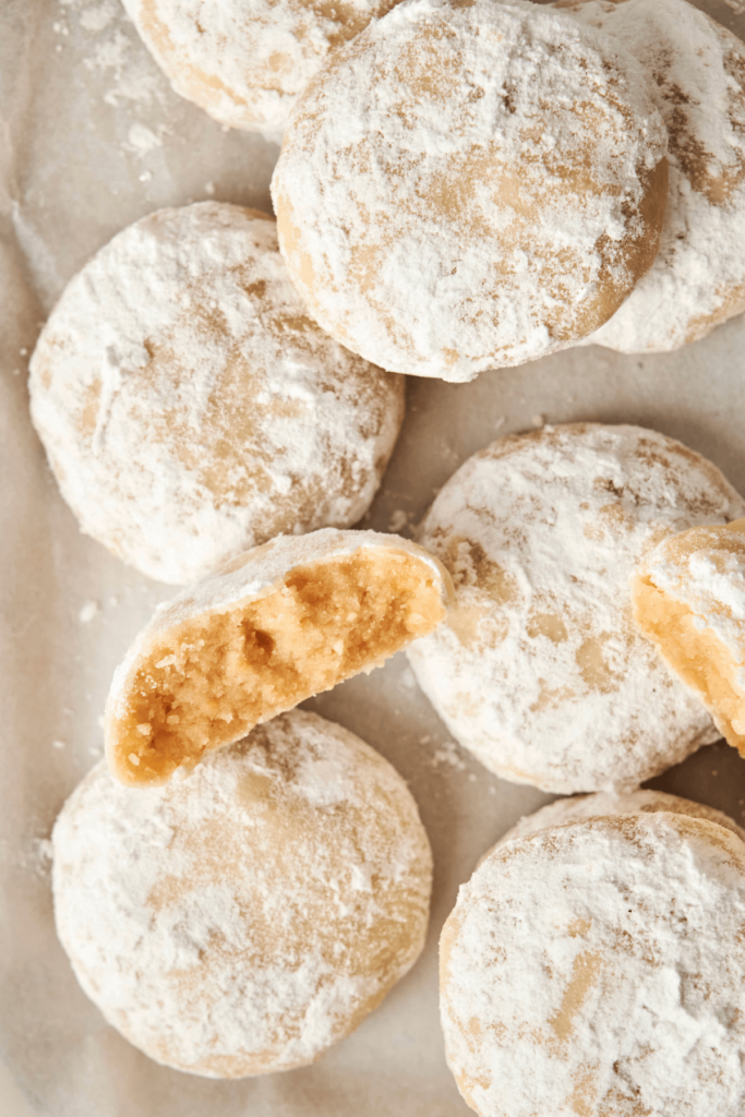 Un primer plano de las Galletas de Almendra en Forma de Nieve cubiertas con azúcar en polvo, con una galleta rota por la mitad para revelar su interior suave y desmenuzable.