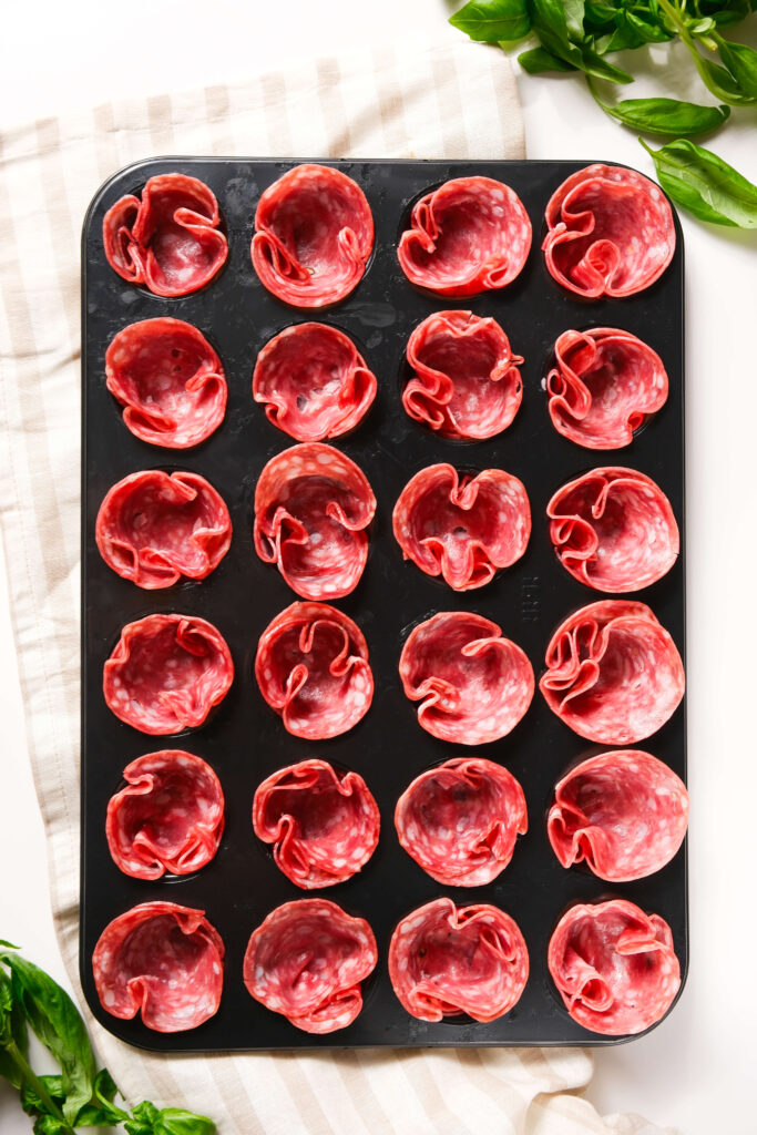 Molde para mini muffins lleno de salami cortado en finas lonchas formando tazas, listo para hornear como bocados crujientes.